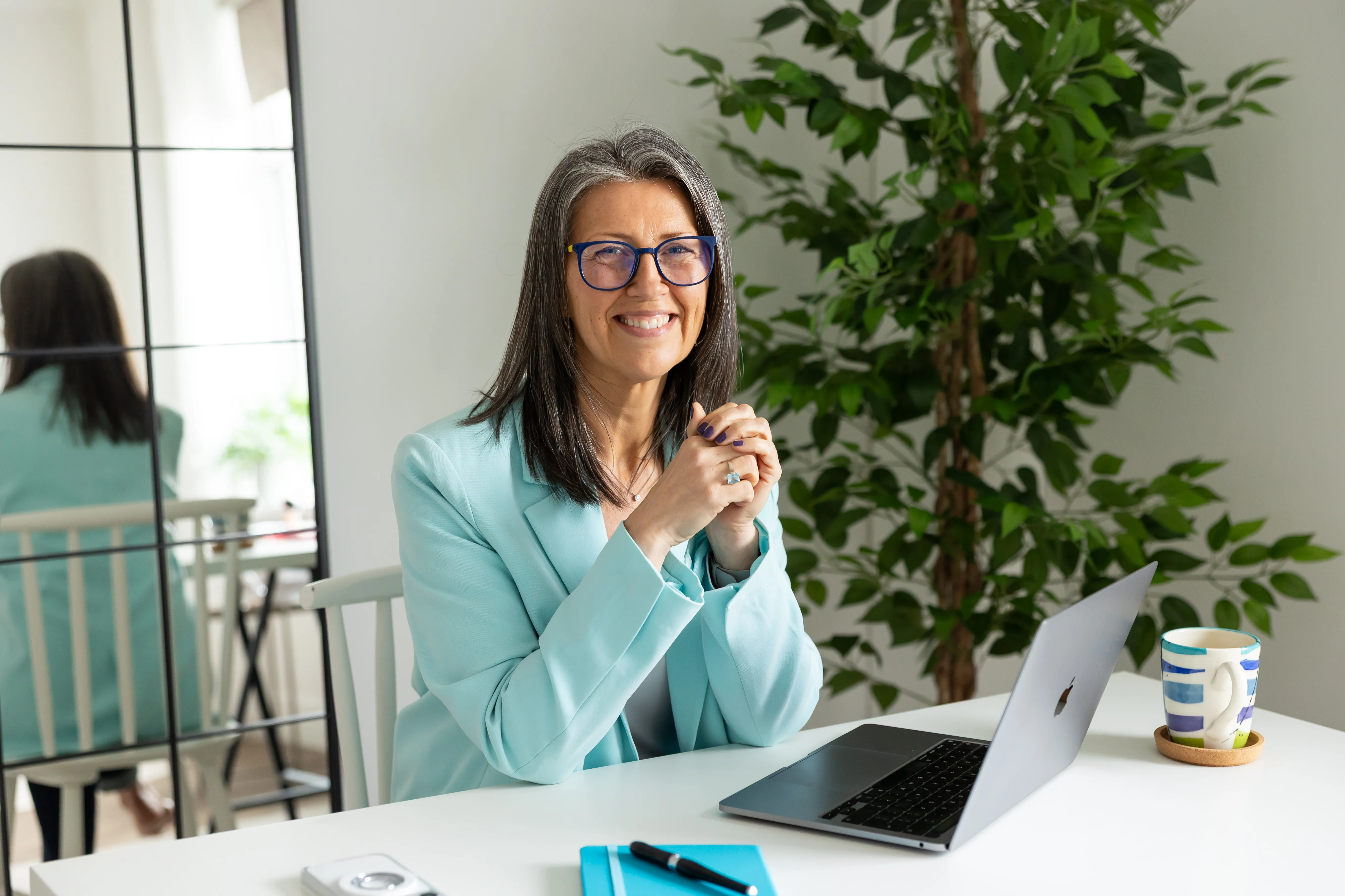 Deborah Bulcock wearing aqua coloured blazer, glasses. Sat at a white desk with laptop and notebook. Large plant standing behind.
